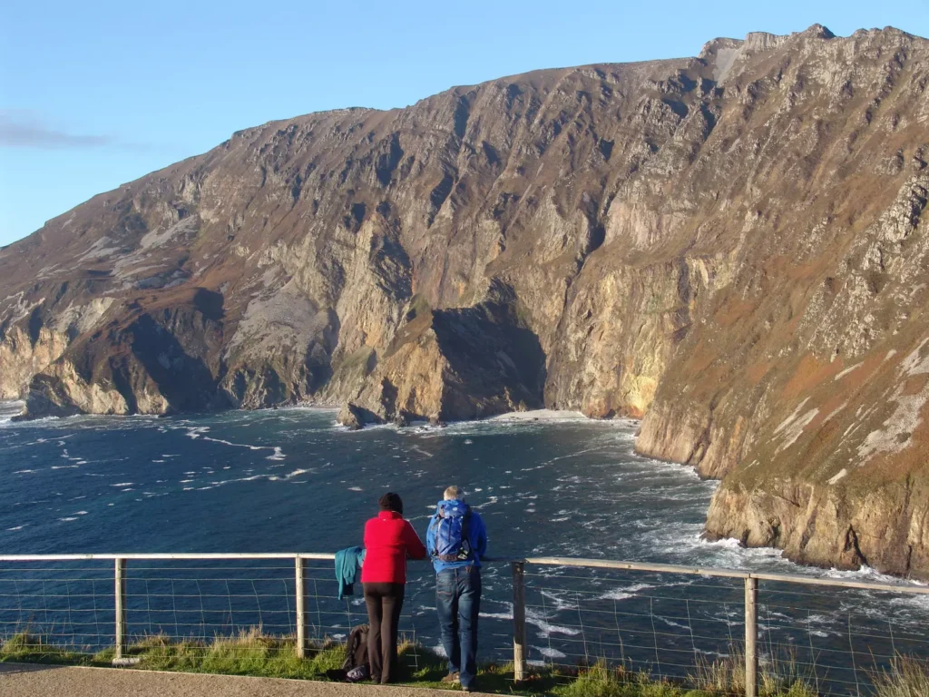 Two hikers leaning on a fence over looking the sea with cliffs in the background.